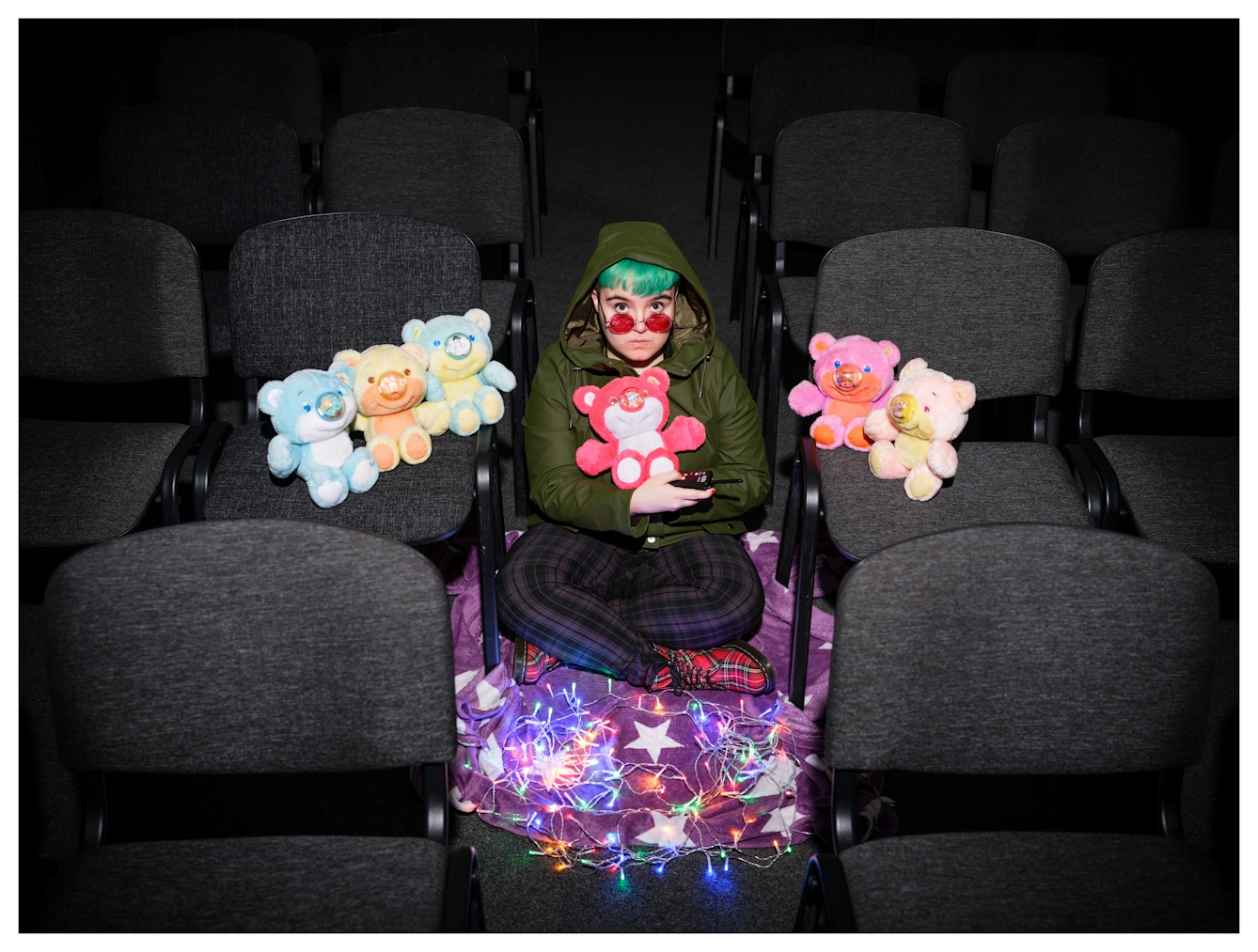 Assisted Self-Portrait of an individual sat crossed legged on the floor of a dark conference room surrounded by grey chairs. They are wearing a green coat with he hood up, have green hair and are wearing rose coloured glasses which they are peering over the top of. In their arms they are cradling a bright red Nosy Bear and on the chairs either side of them are 5 further splashes of colour in the form of other Nosy Bears. They are surrounded by colourful fairy lights and in her right hand is the remote trigger used to capture the image.