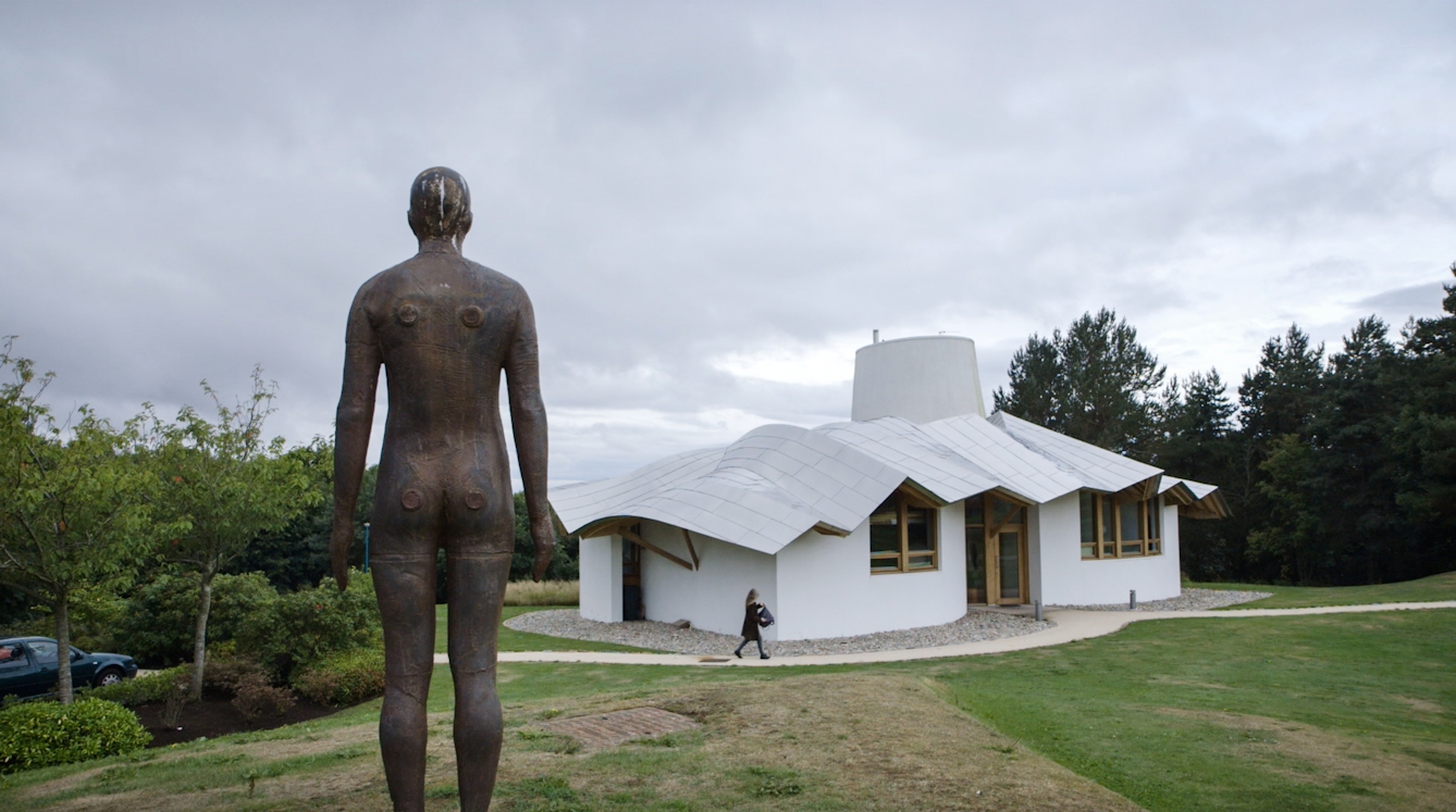 A still from a video about Maggie's Centres, taken outside showing a sculpture of a human facing a small white house.