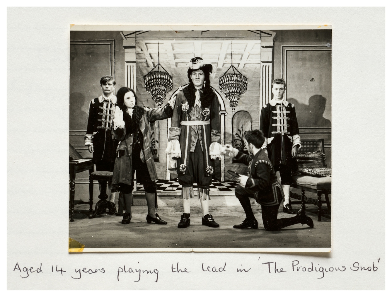 Photograph of a sepia tones photograph from the 1960s stuck into a scrapbook. The photo shows a school production of a play with 5 school children dressed in period costumes on a stage with painted backdrop. In the centre is a tall figure looking straight to camera. Underneath the photograph are the handwritten words '14 years playing the lead in 'The Prodigious Snob'.