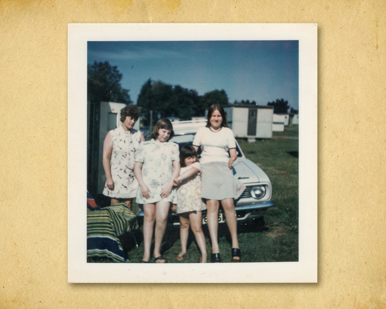 Photograph of a colour photographic print, resting on a brown paper textured background. The print shows a family of 3 daughters and their mother outside on a caravan site. They are leaning on the bonnet of their car, looking to camera, squinting slightly into the sun. 