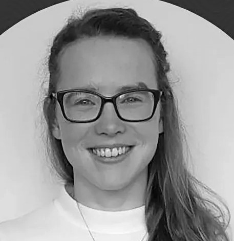 A black and white head-and-shoulders photograph of a young white woman with long fair hair and square framed glasses. She is looking straight at the camera and smiling.