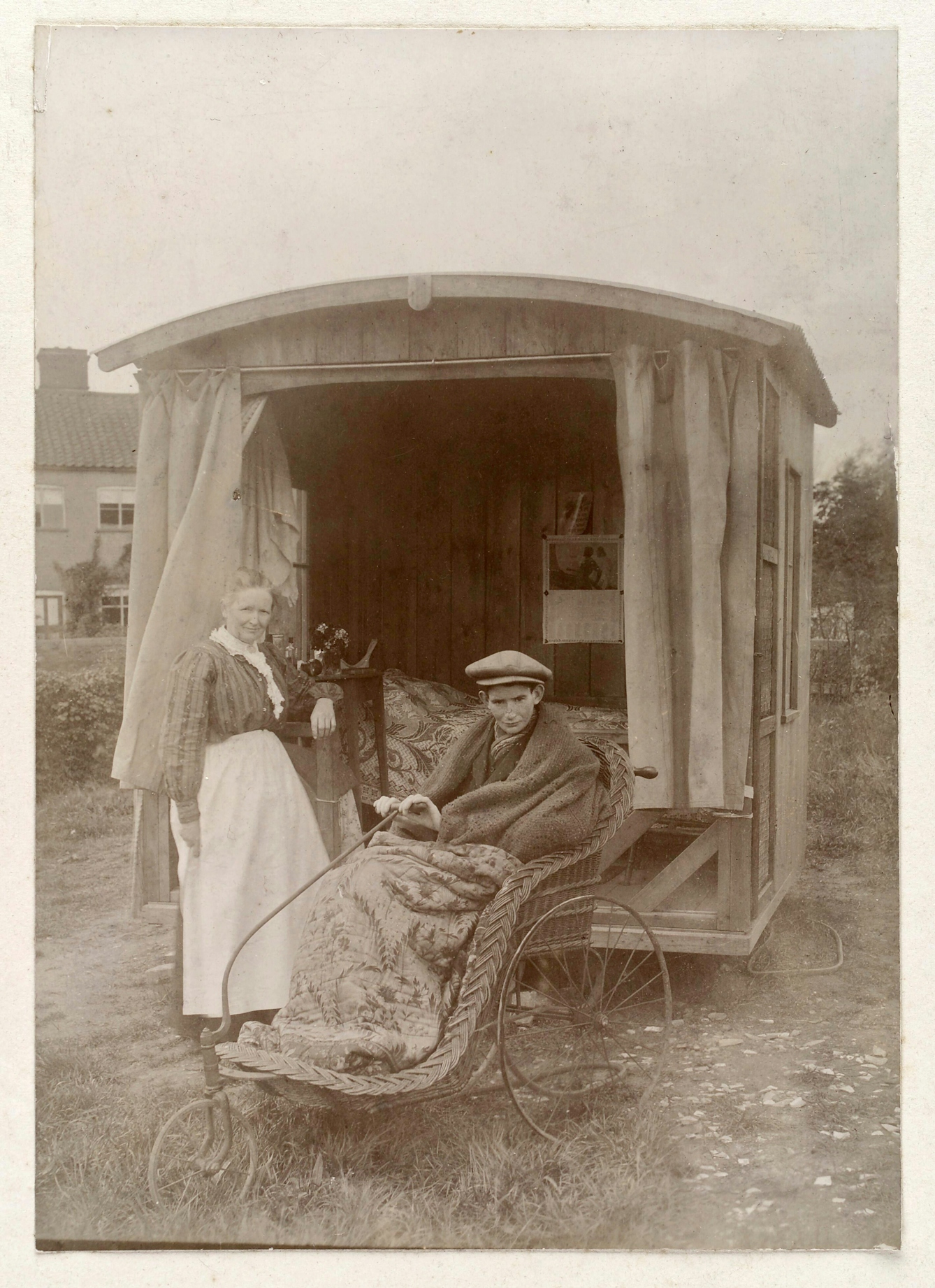 A black and white photograph of an adolescent boy wrapped in blankets and wearing a cloth cap sitting in a whicker bath chair. The chair is infront of a small wooden 'chalet' containing a bed. A nurse in uniform stands to the left of the boy, leaning against the open back of the chalet and both she and the boy gaze directly at the camera.
