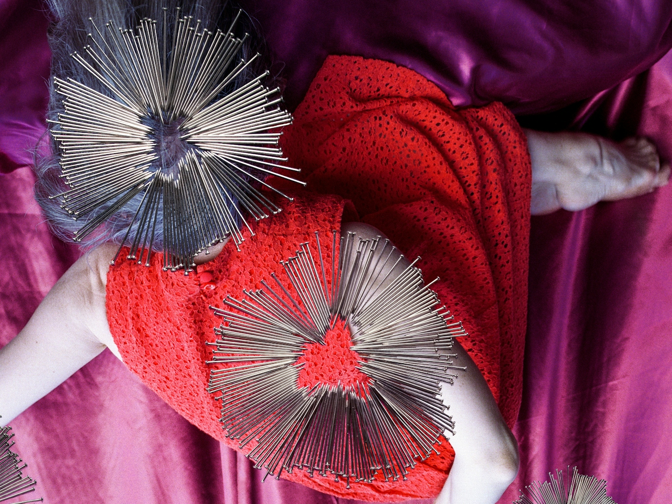 Detail from a larger artwork created with a colour photographic print of a female figure in a bright red dress, set against a purple and blue draped silk background. The figure is on her side, curled up with her knees raised to her chest. Her arms are extended out behind her back. Her body is covered by groups of dress pins, laid on top of the photographic print. The pins are arranged in circles so that the sharp end of the pins point to the centre of the circle. One group covers the figure's head, and another her back.