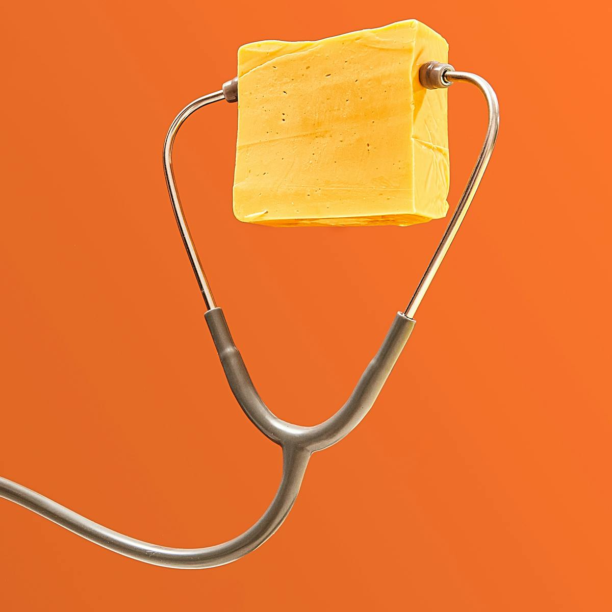 Photograph of a grey and silver stethoscope floating in front of a bright orange background, the tubing curling in a snake-like shape. The chest-piece is just out of frame. Between the earpieces is a large rectangular block of unwrapped yellow cheddar cheese. 