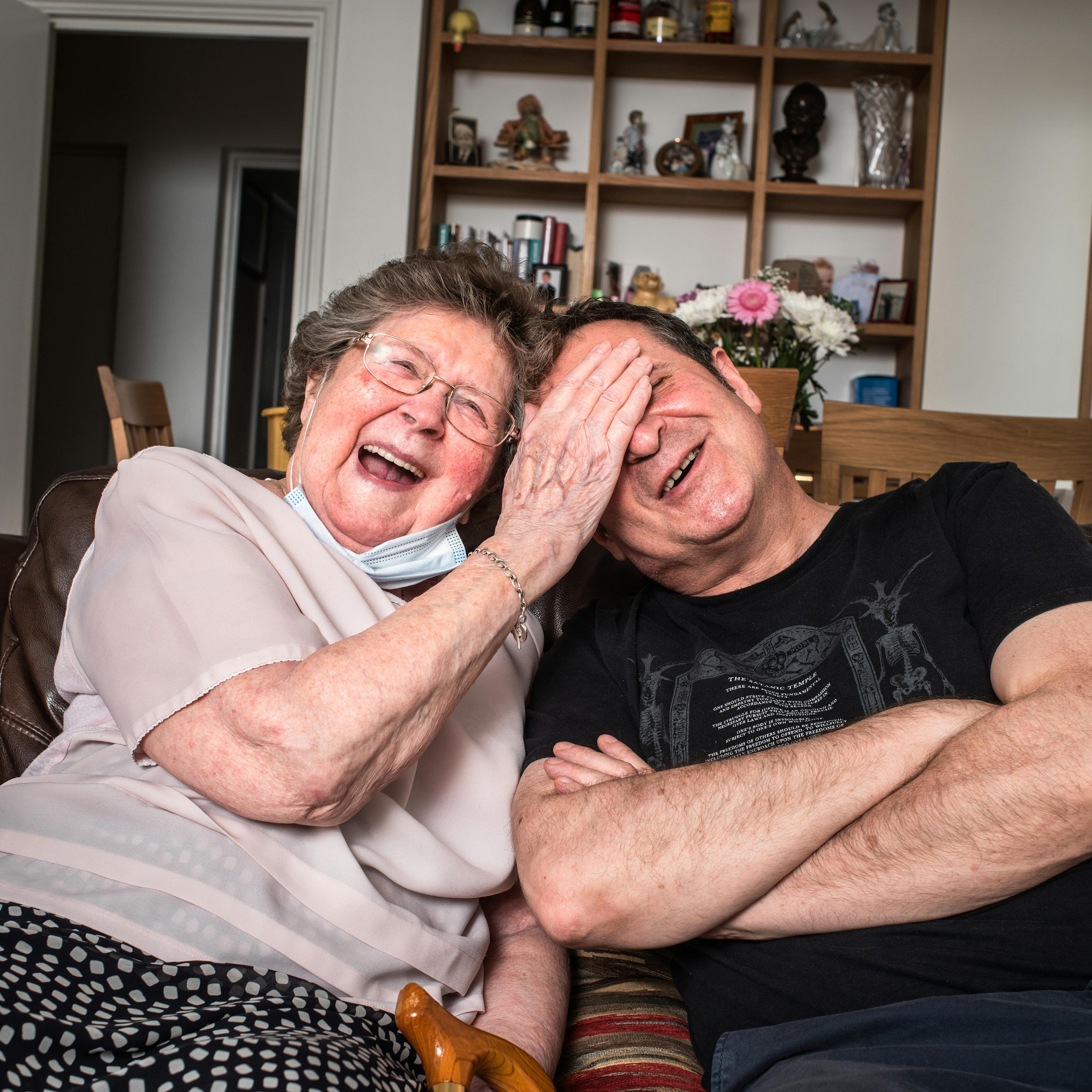 Photograph of a man and his mother sat side by side on a sofa in a living room. The man is seated on the right with his arms crossed. He is leaning towards his mother. His mother seated on the left is laughing out loud and leaning in towards her son, placing her right hand over his eyes and forehead. In the background is a shelving unit containing books, ornaments, bottles of spirits and family photographs.