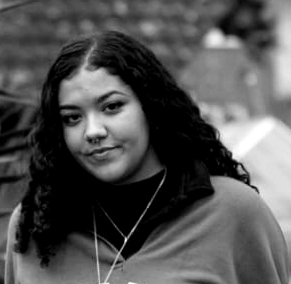 Black and white photograph of the head and shoulders of a young woman with dark shoulder length curly hair. She is looking at the camera and smiling.