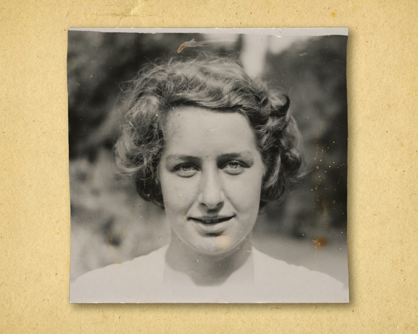 Photograph of a black and white photographic print, resting on a brown paper textured background. The print shows the head and shoulders of a woman outside in a garden. She is looking to camera, wearing a white shirt.