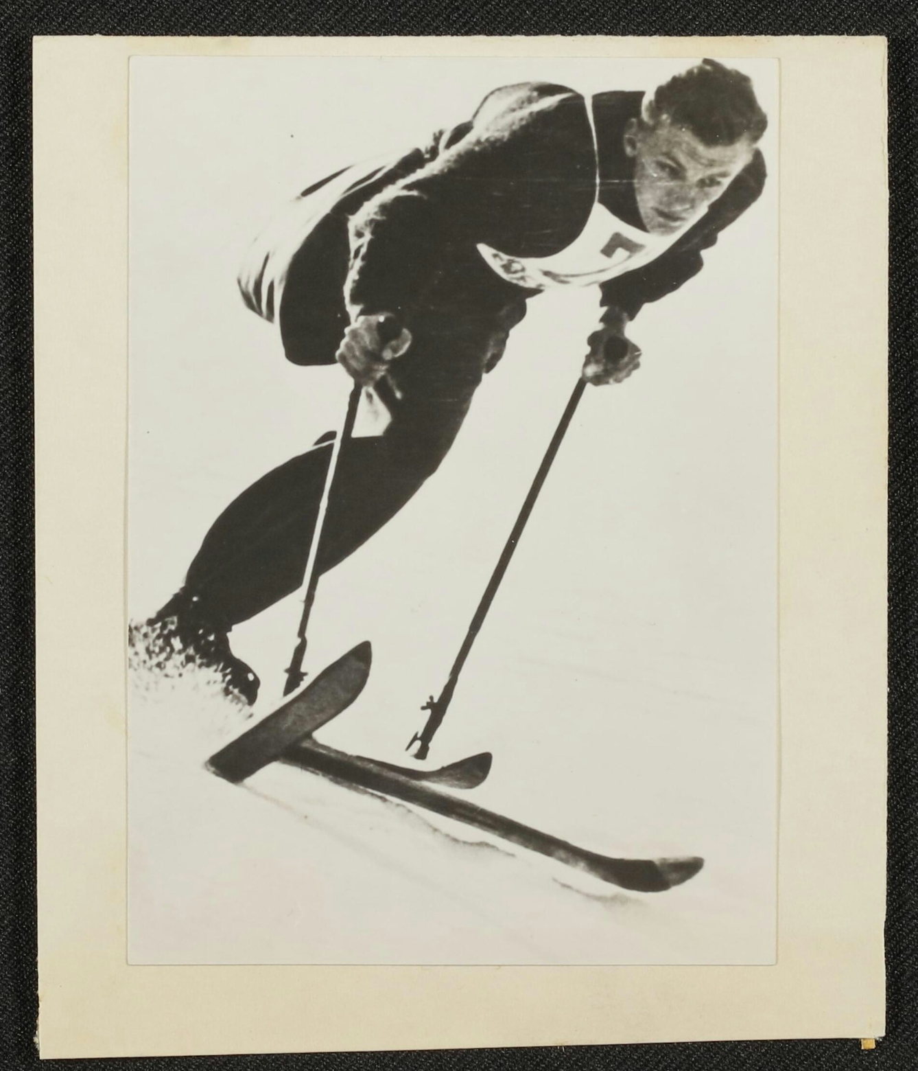 Black and white photographic print of a man skiing. He has one leg and one stump. He leans forward, gripping his poles, and snow sprays up from under his ski, indicating speed. His expression indicates concentration as he gazes off toward the bottom right of the image.