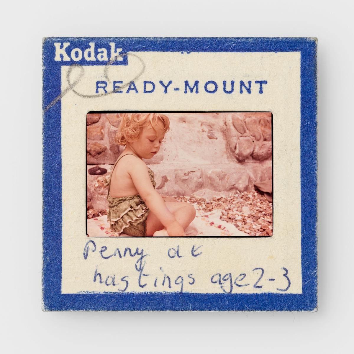 Photograph of a colour 35mm transparency mounted in a cardboard side holder, resting on a white background. The transparency shows a young girl in a frilly green bathing suit sitting on a towel on a shingle beach, by a stone wall. The slide mount has a blue border around the edge with Kodak Ready-mount printed on it. A hand written note on the mount reads, "Penny at Hastings age 2-3".