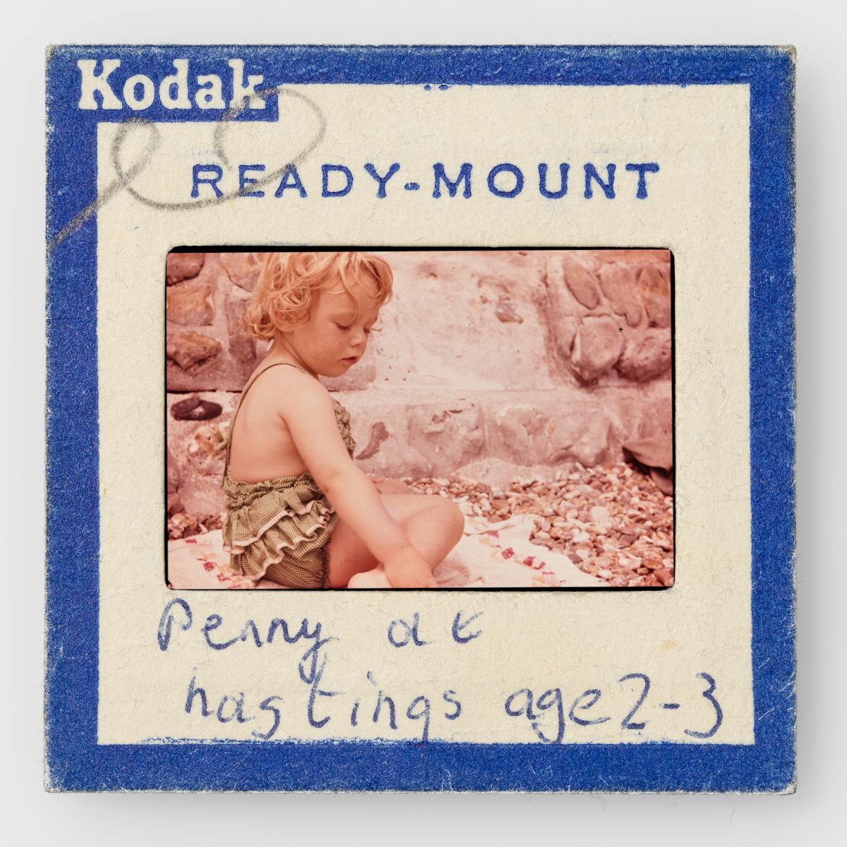 Photograph of a colour 35mm transparency mounted in a cardboard side holder, resting on a white background. The transparency shows a young girl in a frilly green bathing suit sitting on a towel on a shingle beach, by a stone wall. The slide mount has a blue border around the edge with Kodak Ready-mount printed on it. A hand written note on the mount reads, "Penny at Hastings age 2-3".