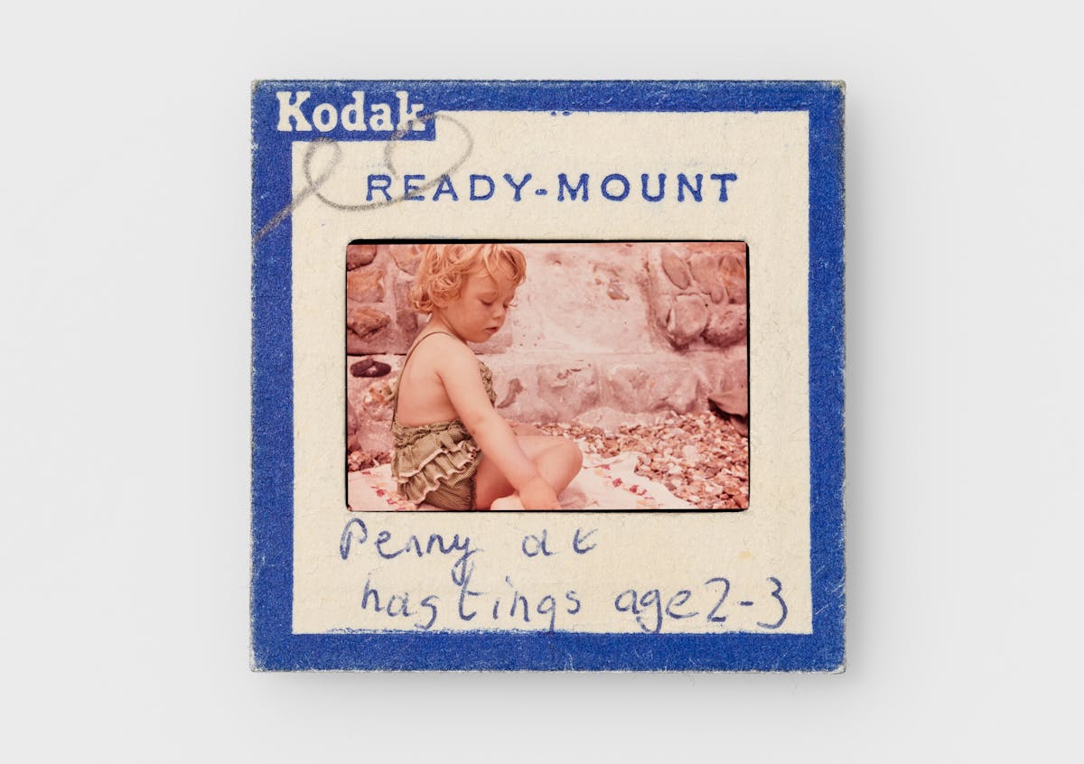 Photograph of a colour 35mm transparency mounted in a cardboard side holder, resting on a white background. The transparency shows a young girl in a frilly green bathing suit sitting on a towel on a shingle beach, by a stone wall. The slide mount has a blue border around the edge with Kodak Ready-mount printed on it. A hand written note on the mount reads, "Penny at Hastings age 2-3".