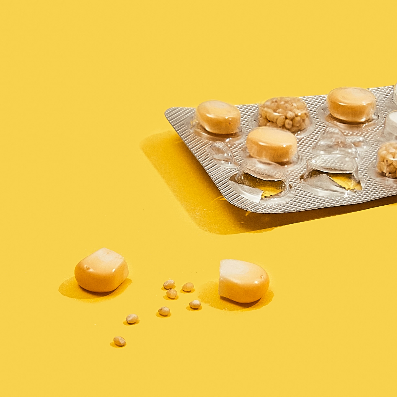 Photograph of a foil medicine pill blister pack sitting on a bright yellow background. The blisters of the pack contain either a single kernel of corn or a collection of small grains of maize. A couple of the blisters are empty and there are 2 kernels of corn and a scattering of maize grains spread out on the yellow background.