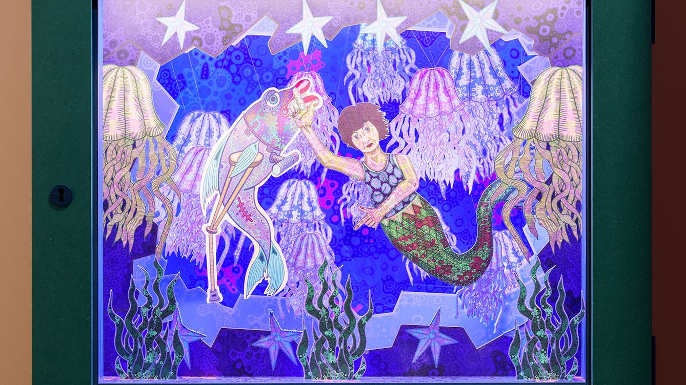 Photograph of a diorama called Mum the Mermaid. A mermaid is swimming with fish and jellyfish under water and she is pointing up with one arm and pointing down with the other arm.