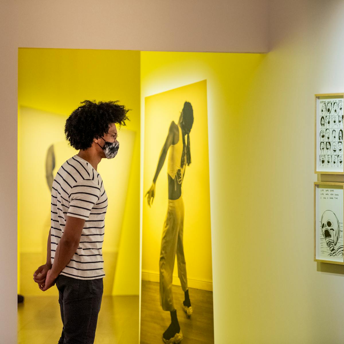 Photograph of a man wearing a face mask exploring an exhibition gallery space. He is wearing a white t-shirt with thin black horizontal stripes and jeans. His hands are clasped behind his back. The gallery around him has a warm yellow and pink tone. Directly in front of him is a large photographic print of a person in full length, frozen in a performative pose. Hung on the wall to the right is a cluster of 6 framed pen and ink drawings, 3 across by 2 high, showing in one a skull, in another a bird cage and in another a series of hands.