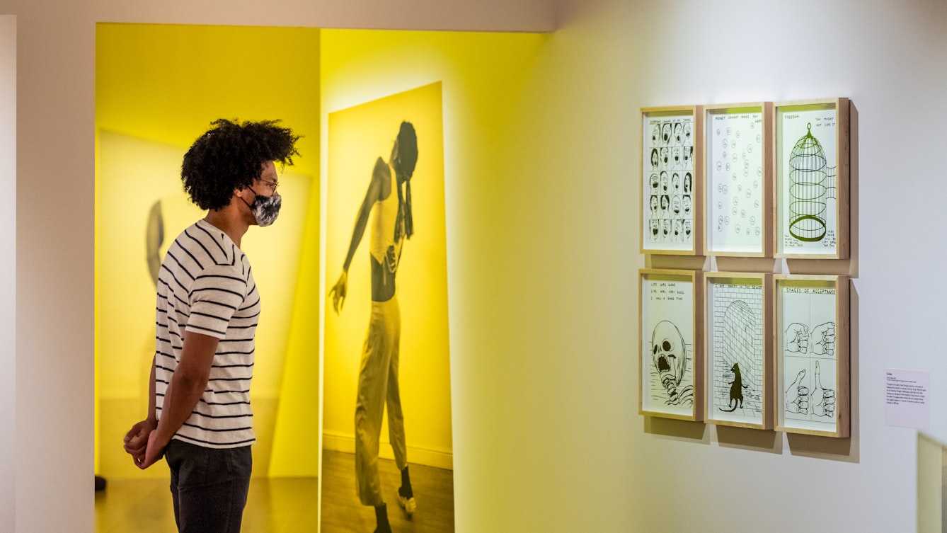 Photograph of a man wearing a face mask exploring an exhibition gallery space. He is wearing a white t-shirt with thin black horizontal stripes and jeans. His hands are clasped behind his back. The gallery around him has a warm yellow and pink tone. Directly in front of him is a large photographic print of a person in full length, frozen in a performative pose. Hung on the wall to the right is a cluster of 6 framed pen and ink drawings, 3 across by 2 high, showing in one a skull, in another a bird cage and in another a series of hands.