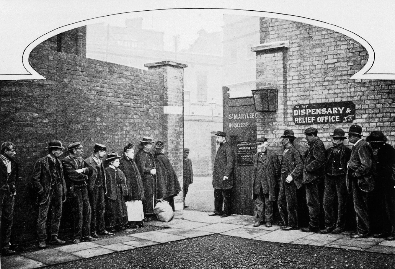 Black and white photograph showing people queuing outside Marylebone workhouse. Men make up the majority of the queue, although there are a few women. There is a sign behind one portion of the queues that reads 'Dispensary relief office'. 