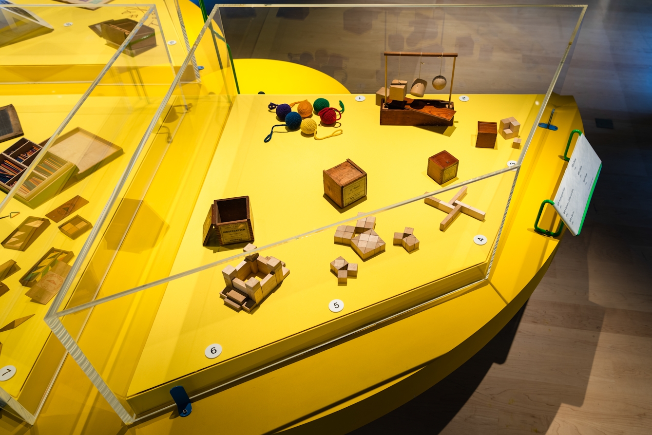 Photograph of an exhibition display case with a bright yellow base. Inside the case are several small wooden toys in various shapes and 6 balls of coloured wool.