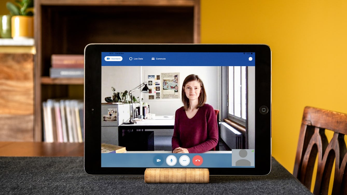Meekyung MacMurdie appearing on a video call on a tablet device. Meekyung is sitting at a desk wearing a red top with mid-length brown hair. The tablet is appearing propped up on a wooden table with a chair and bookshelves and a yellow wall in the background.