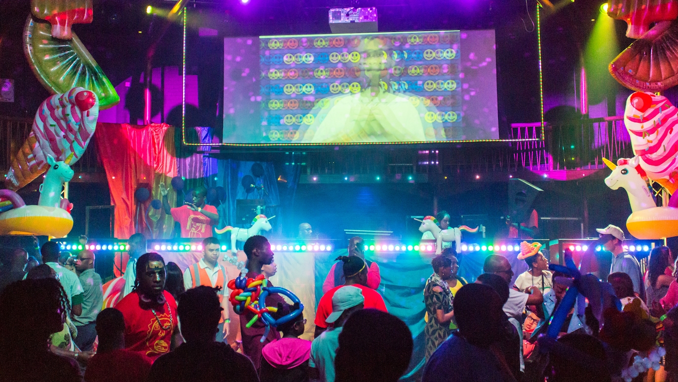 Photograph of the Beautiful Octopus Club. At the club night people are wearing vibrant, colourful clothes, there are bright lights, balloons, inflatable unicorns and DJs on stage. 