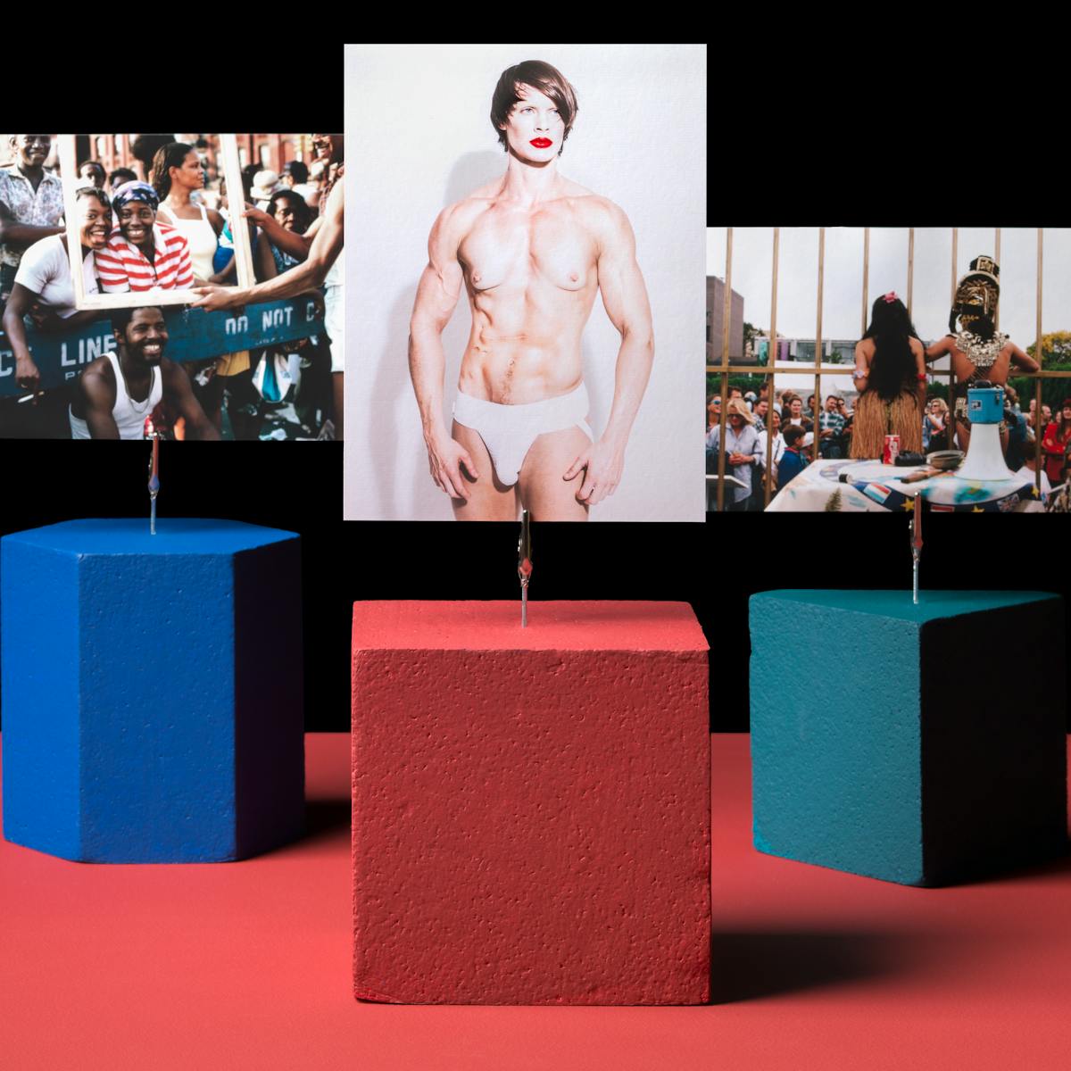 Photograph of three shapes holding three images on a red tabletop with crocodile clips.  The red cube is holding a portrait image of a largely naked person with red lipstick and white thong.  The teal triangle is holding a picture of a performance where two people, a man and a woman, are in costume with their backs to the camera whilst addressing a crowd.  A blue hexagon is holding a picture of a crowd of people standing over a ‘do not cross’ barrier.  The mood of the crowd is joyous.  Two women are posing to the camera with beaming smiles whilst a picture frame is being held in front of them by another person.  The picture frame is also resting on the head of a fourth person.
