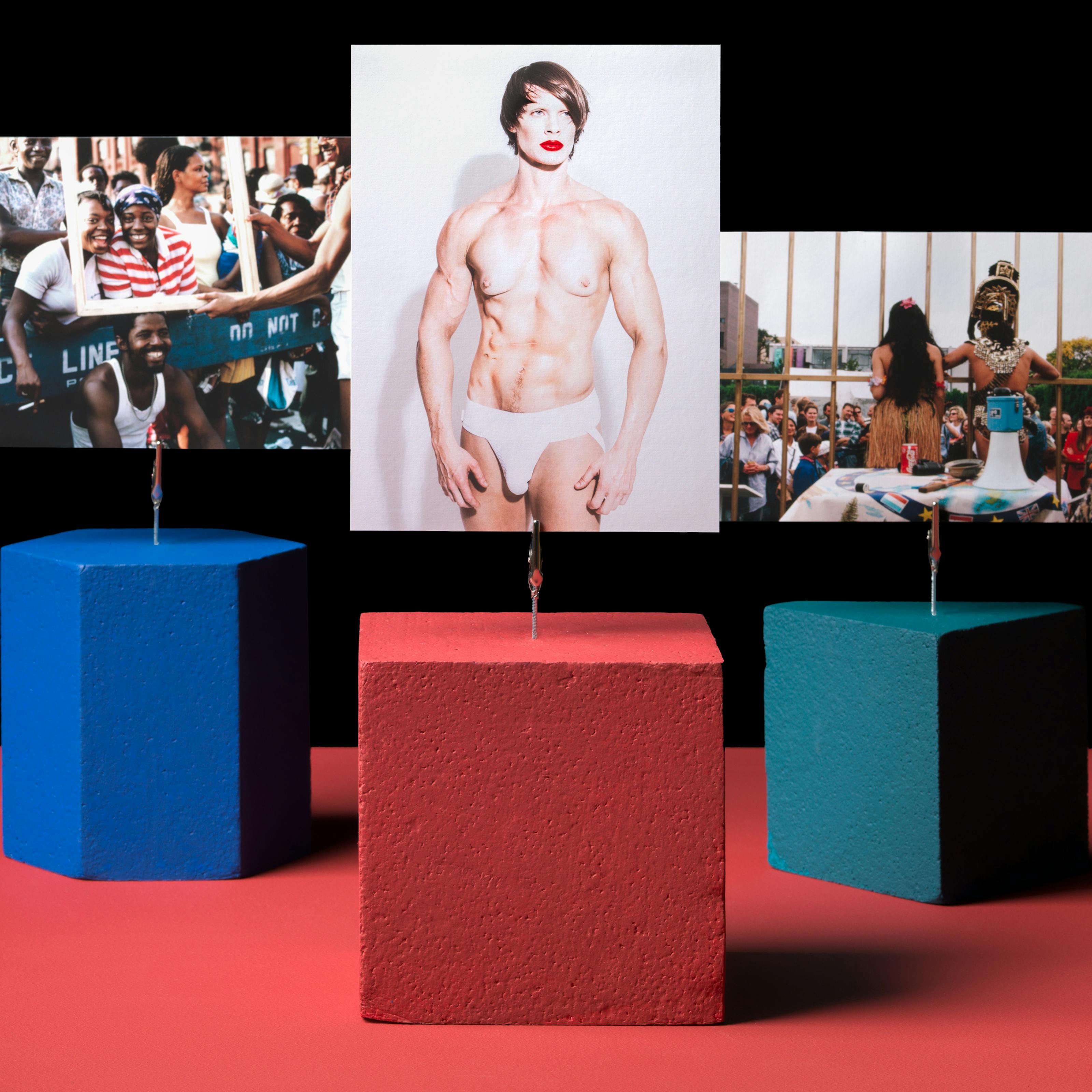 Photograph of three shapes holding three images on a red tabletop with crocodile clips. The red cube is holding a portrait image of a largely naked person with red lipstick and white thong. The teal triangle is holding a picture of a performance where two people, a man and a woman, are in costume with their backs to the camera whilst addressing a crowd. A blue hexagon is holding a picture of a crowd of people standing over a ‘do not cross’ barrier. The mood of the crowd is joyous. Two women are posing to the camera with beaming smiles whilst a picture frame is being held in front of them by another person. The picture frame is also resting on the head of a fourth person.