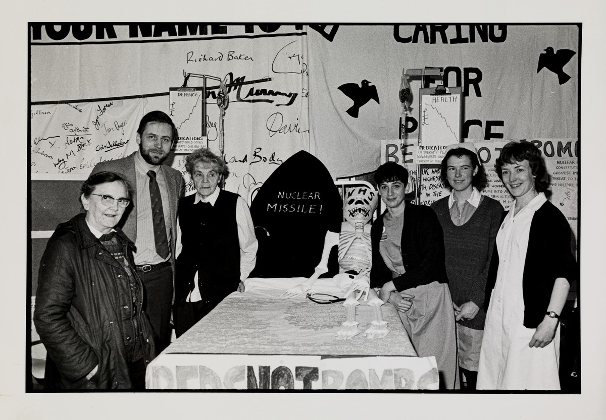 A black and white image showing 5 women and a man stood round a table, surrounded by nuclear protest posters.