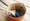 Photograph of a well worn cast iron and enamel coated saucepan with a wooden handle, standing on a table top made of worn wooden planks. In the saucepan are blackberries with boiling liquid being poured over them. A wooden spoon stirs the mixture.