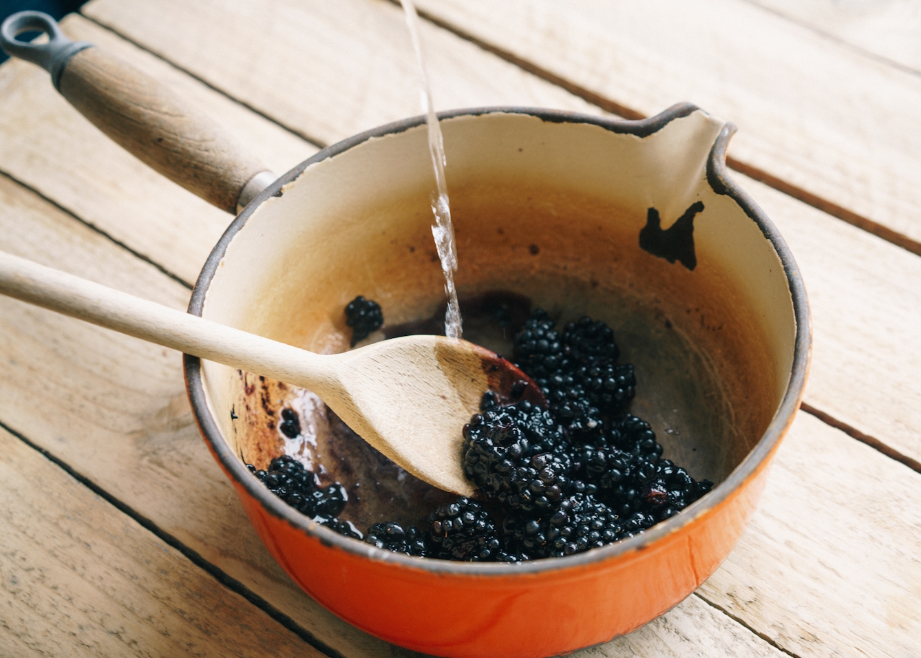 Photograph of a well worn cast iron and enamel coated saucepan with a wooden handle, standing on a table top made of worn wooden planks. In the saucepan are blackberries with boiling liquid being poured over them. A wooden spoon stirs the mixture.