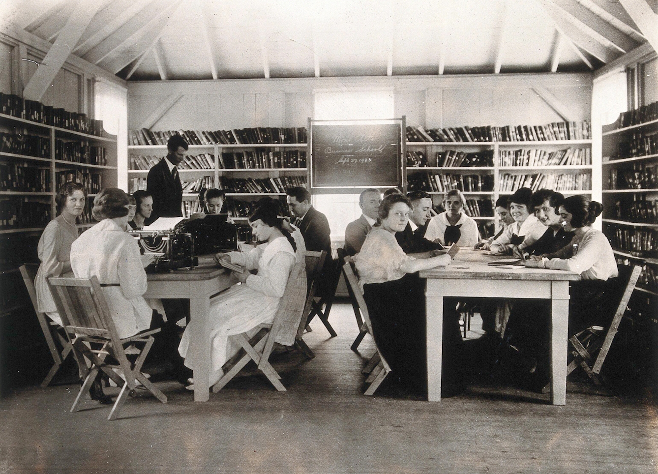 Black and white photograph of the interior of a room lined with bookshelves and a blackboard. Seated around the two large tables in the room are lots of people with books and typewriters, some looking up at the camera.