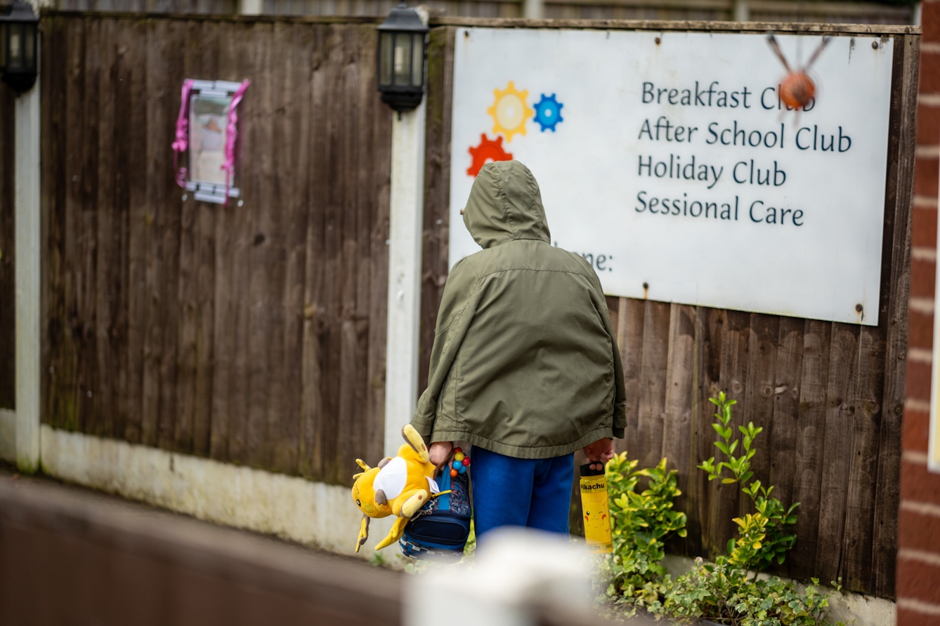 A photograph of a child carrying stuffed toys and a water bottle, at the entrance of a school which is advertising after school and holiday clubs.