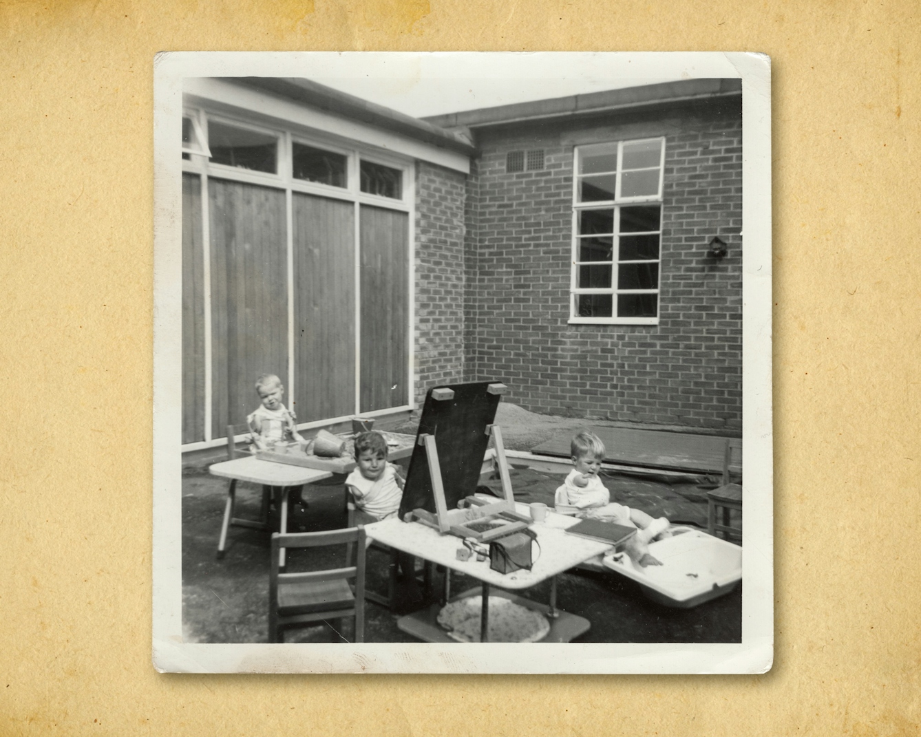 Photograph of a black and white photographic print, resting on a brown paper textured background. The print shows a group of 3 young children seated outside a school building, all engaged in an activity of drawing on a blackboard, playing in a sandpit or with a water. All the children have been affected by the thalidomide scandal and so some have short arms, some have short legs, and some have both.