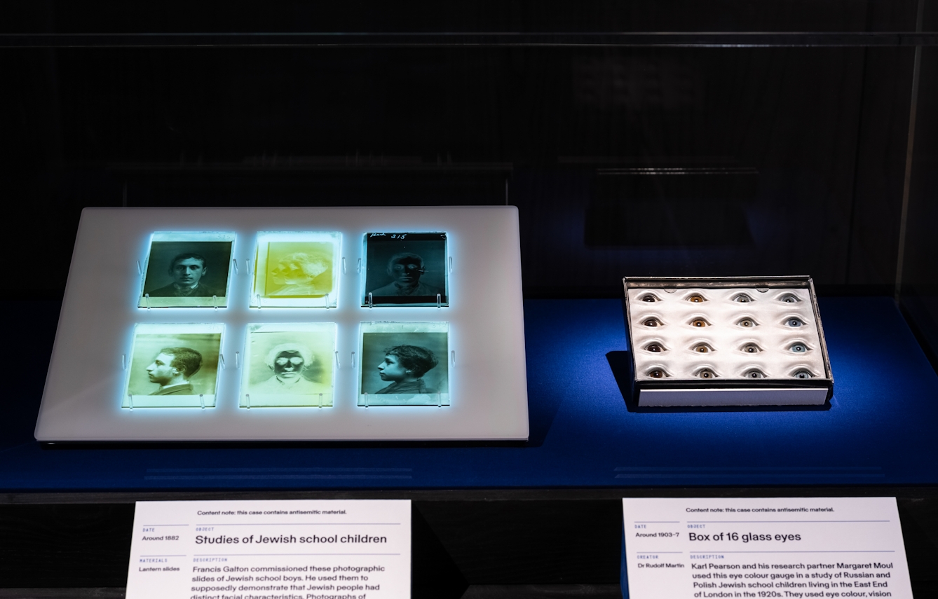 Photograph of an exhibition display case in a dark gallery space. The case contains 2 exhibits. On the left is a series of 6 photographic negatives and transparencies in a 3 by 2 grid, backlight on a lightbox. On the right is a box of glass eyes containing 16 eyes in a. 4 by 4 grid. In front of the case are 2 information panels.