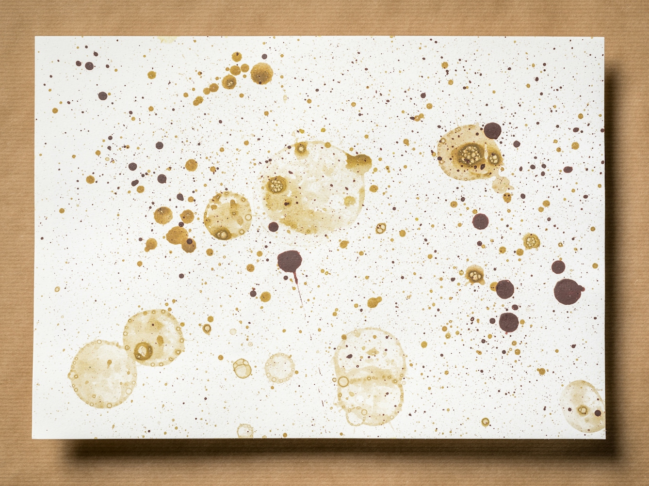 Photograph of an original artwork on watercolour paper. The artwork is resting on a brown parcel paper background. The artwork shows many varying in size droplet spatters and rings, made with coloured ink. The ink colours are dark and light greens. The spatters seem random in distribution.