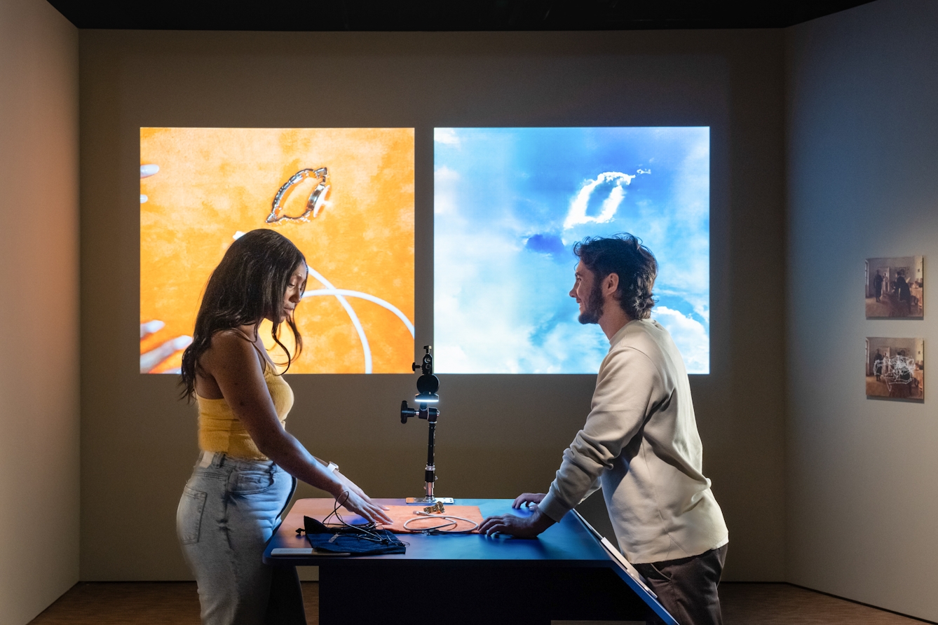 Photograph of two people exploring an exhibition. They are stood facing each other over a tabletop. On the table are a selection of object which they are arranging under a camera. Behind them on the gallery wall there are two square projections. On the left is the camera image of the objects they have arranged, a watch, cables against an orange background. On the right is a computer generated interpretation of their arrangement, abstract white shapes on a blue background.
