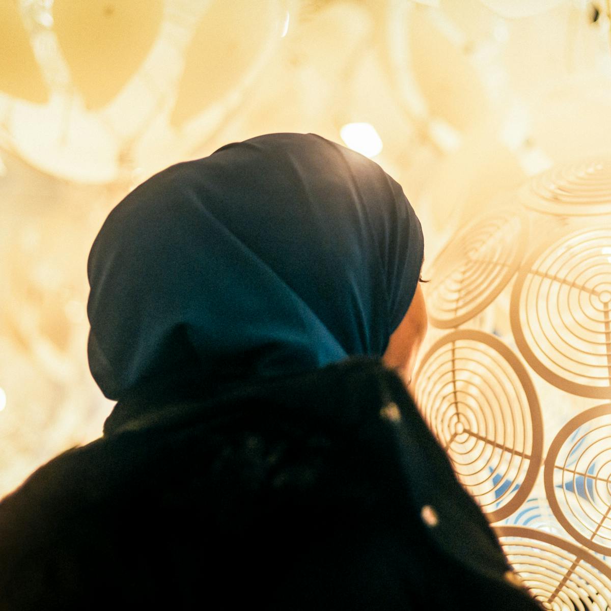 Photograph of a woman wearing a blue head scarf facing away from the camera. The viewpoint is low down looking up at her. In the background is the warm yellow and orange colours of patterned lamp shades in a shop display.