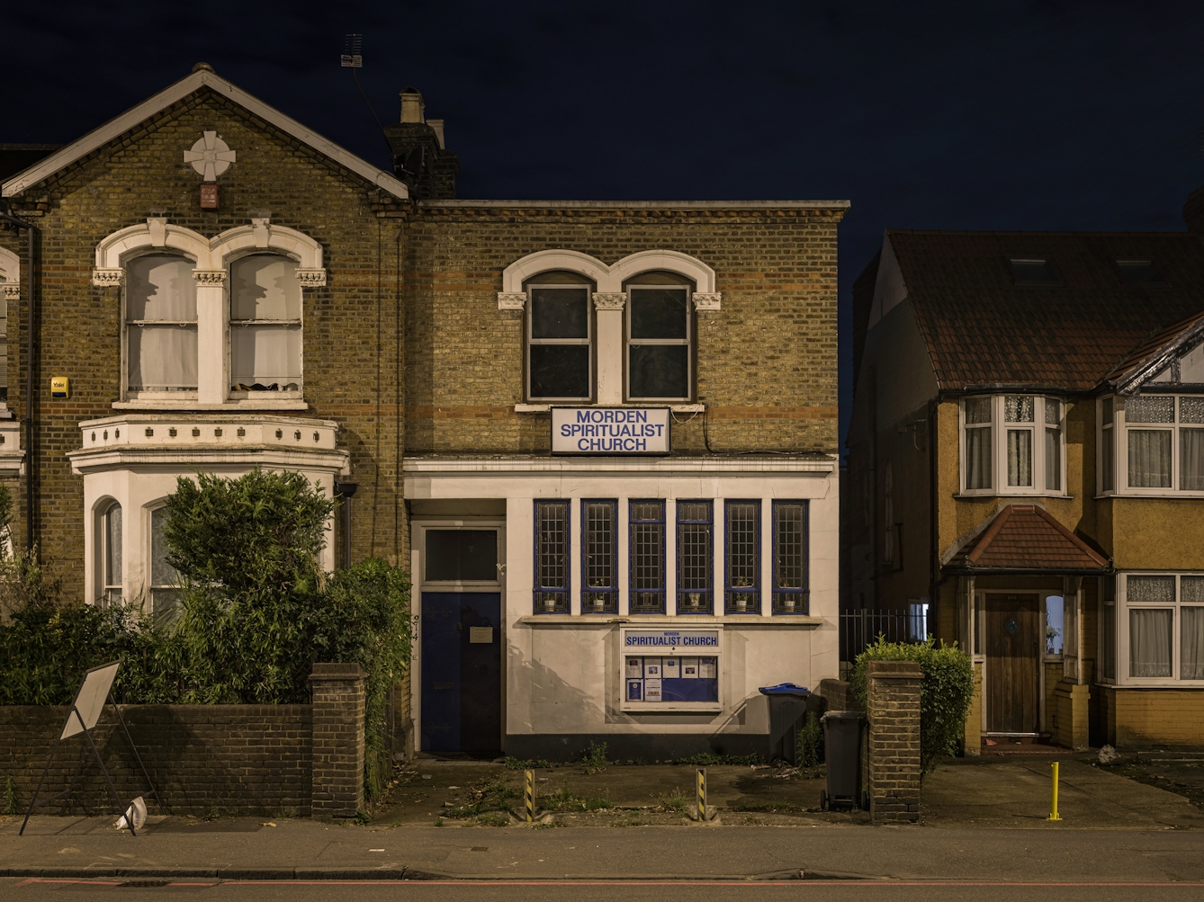 Photograph of Morden spiritualist church at night.  The building is uncharacteristic of typical church architecture, and appears to be from the Edwardian period.  The bottom half of the building is rendered white, whilst the top half is bare brick.  On the ground floor, the blue door is offset to the left with six tall long windows to the right. A sign with blue letters on white spells 'Morden Spiritualist Church'.