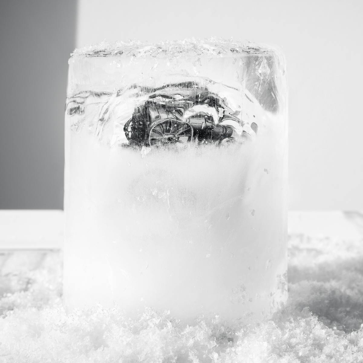 Monotone photograph showing a cylindrical core of frozen water. Encased within the ice is an illustration of a traction steam engine from the 1800s that can just be seen through the distortions of the ice wall and opaque frosting. The ice core is standing vertically on fridge freezer shelf, surrounded by a piles of light frosted snow.