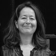 Black and white, head and shoulder photograph of a woman with shoulder length dark hair. She is wearing a patterned shirt and standing in front of a black background.