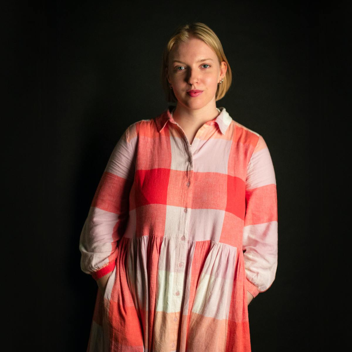 Photographic portrait of a white woman with bobbed blond hair, wearing a red checked dress, standing against a black background. She has her hands in her pockets and quietly smiling straight to camera.