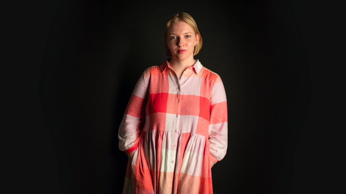 Photographic portrait of a white woman with bobbed blond hair, wearing a red checked dress, standing against a black background. She has her hands in her pockets and quietly smiling straight to camera.