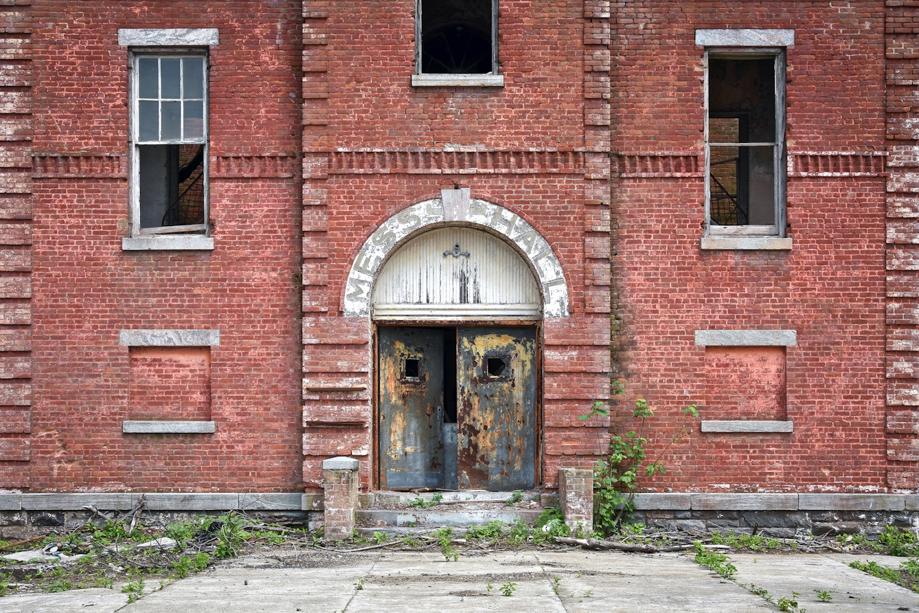 Photograph of the front of an abandoned red brick building, which fills the frame. In the centre is the main door which is slightly ajar. Above the door are the faded letters which spell out 'Mess Hall'. The windows to the left and right above the door are broken and missing their glass.