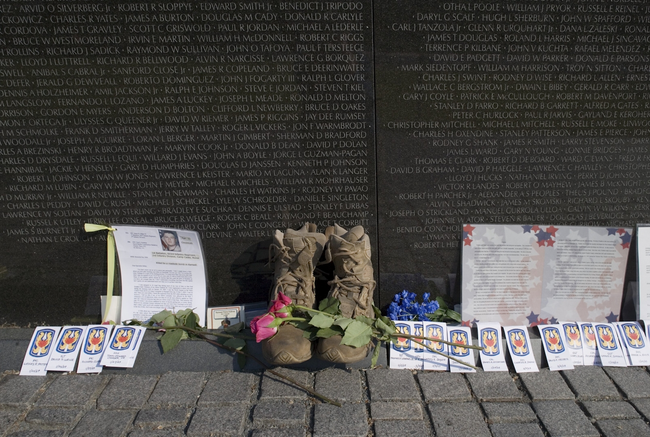 Photograph of objects left at the Vietnam War memorial in Washington. The image shows several obituaries, flowers, badges and a pair of boots which have been left against the memorial. On the marble memorial, engraved white text is visible showing a list of names. 