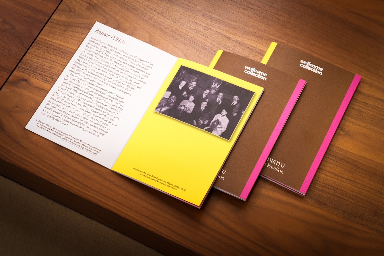 Photograph of three exhibition printed guides resting on a wooden tabletop. The top copy is open at a page with text on the left hand page titled 'Repair' and a black and white archive photograph on a yellow background on the right hand page.