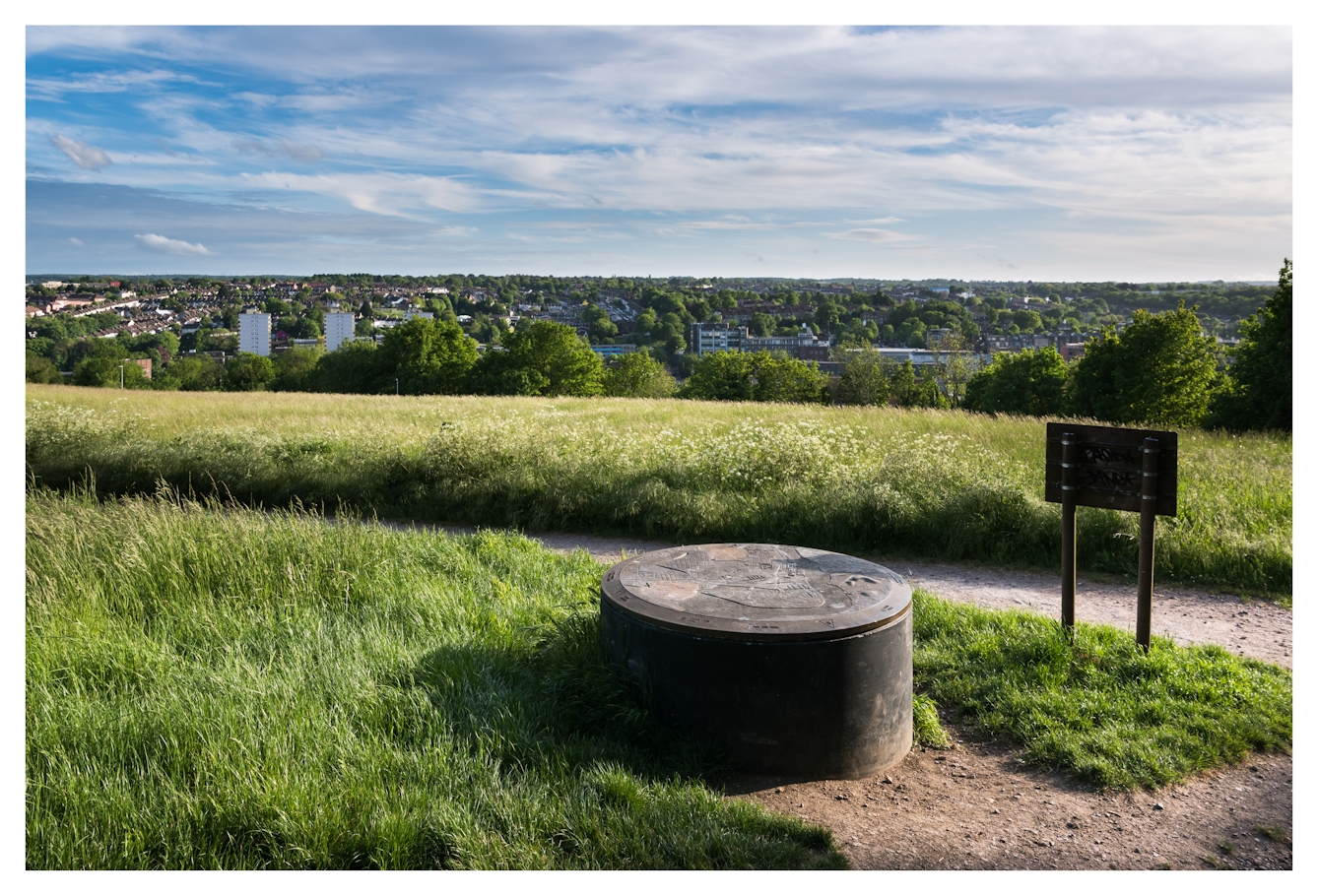 Colour photograph of a hill top expanse of long grassland in a country park setting. In the distance the roofs and tower blocks of a town can be seen. In the foreground, at an intersection of footpaths paths is a low cylindrical concrete structure. On the top surface is a relief maps showing the location and shape of the local towns and topographical landscape.