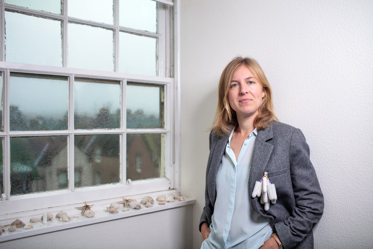 Photograph of a woman next to a window which is covered over the outside in rain drops. On the window sill are an assortment of seashells. The woman is looking straight to camera, wearing a grey jacket.  Hanging from her jacket pocket are about 10 tampons.