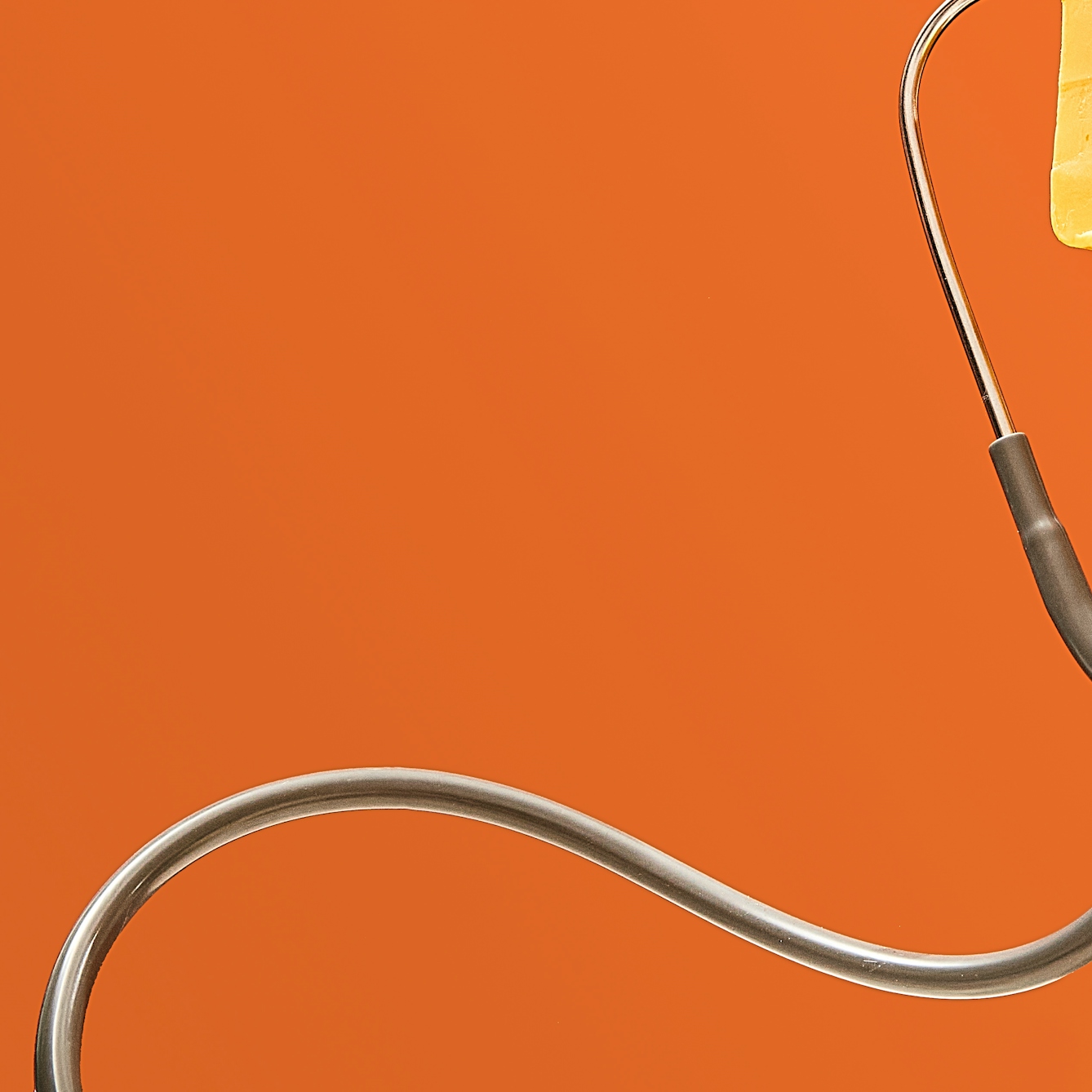 Photograph of a stethoscope floating in front of a bright orange background, the tubing curling in a snake-like shape. The chest-piece is just out of frame. Just visible in the top right corner is the edge of a rectangular block of unwrapped yellow cheddar cheese. 