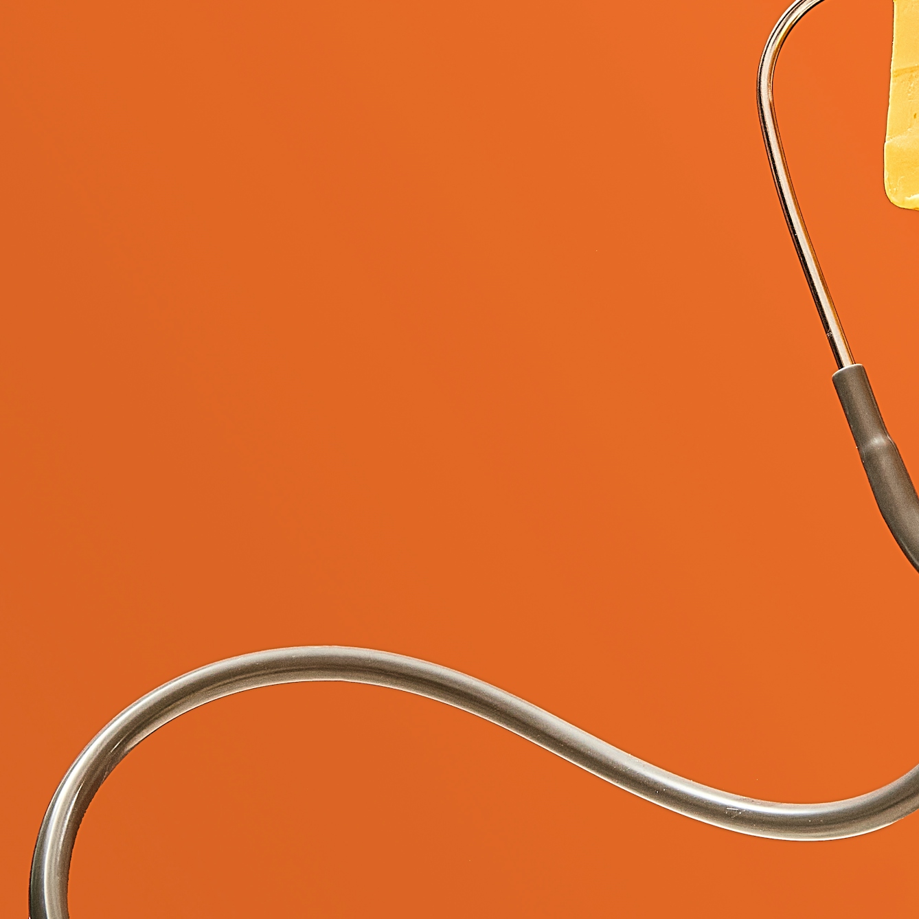 Photograph of a stethoscope floating in front of a bright orange background, the tubing curling in a snake-like shape. The chest-piece is just out of frame. Just visible in the top right corner is the edge of a rectangular block of unwrapped yellow cheddar cheese.