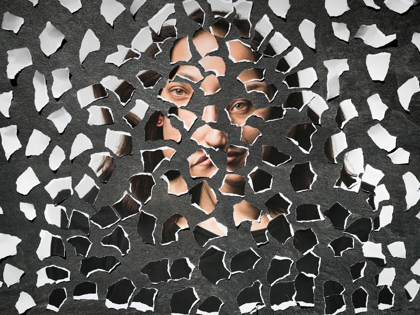 Photograph of a collages of torn up pieces of a photographic print scattered on a grey slate background. There are fragments of eyes, lips, hair and dark and light tones.