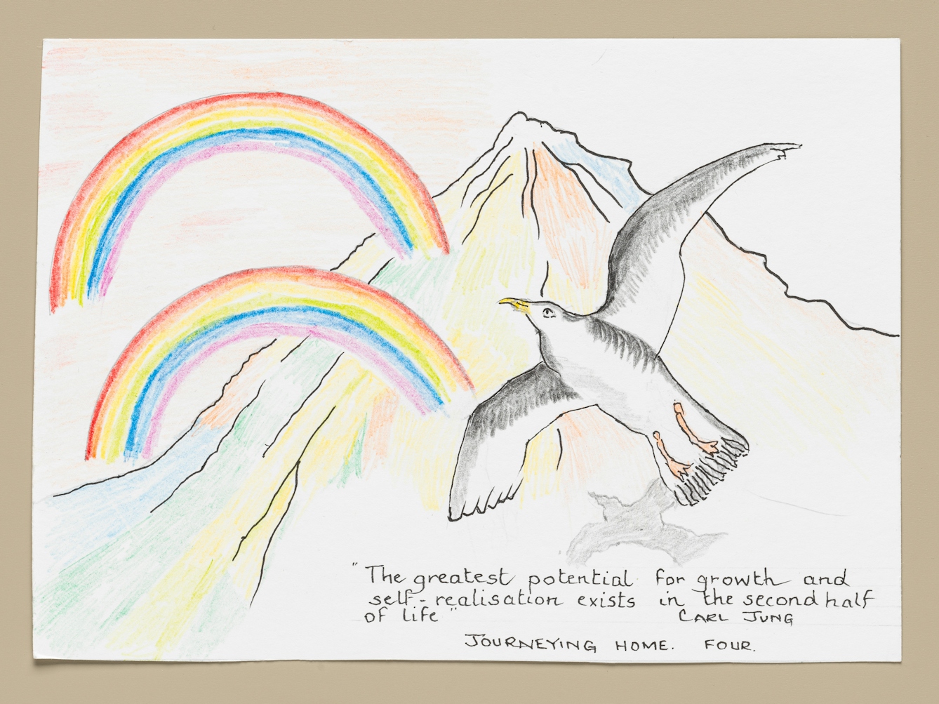 Photograph of an artwork resting on a light taupe-coloured background. The artwork is coloured pencil and ink on textured white paper. It shows a large seagull soaring in the air above a large mountain. In the distance are two large rainbows reaching across half the mountain. The mountain is bathed in rainbow light and colours. Below the scene are the words, “The greatest potential for growth and self-realisation exists in the second half. Carl Jung.”