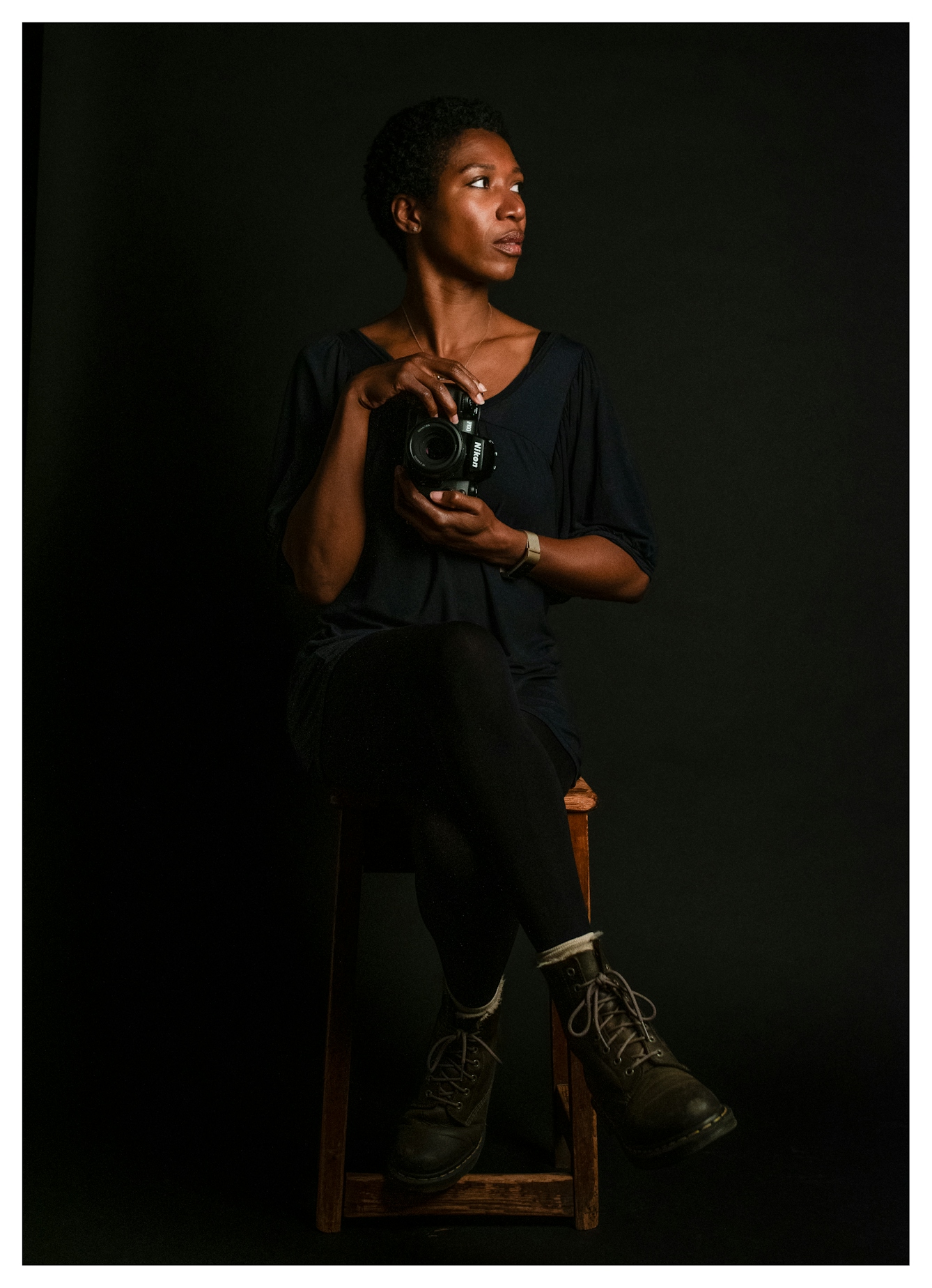 Photographic portrait of a Black woman holding a camera, sitting on a wooden stood against a black background. She is looking away to her left with the air of quite confidence.
